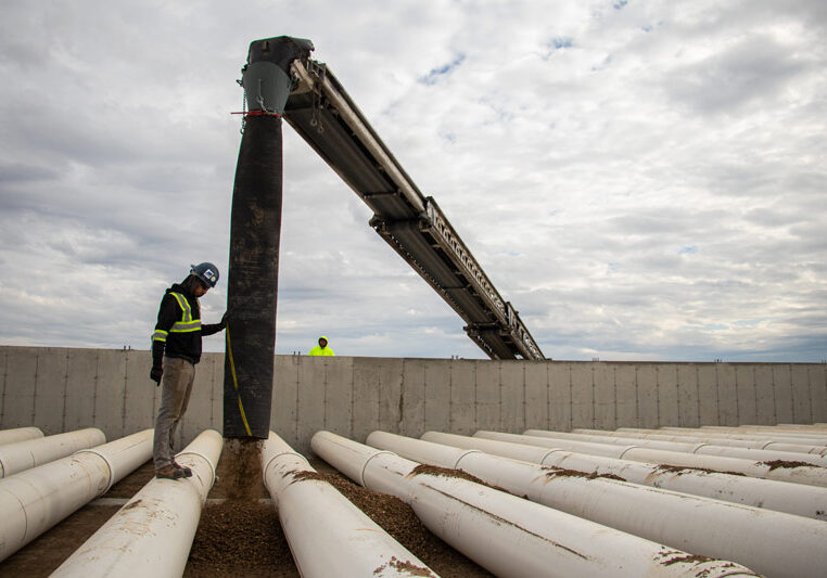 A construction worker guides a telebelt conveyor as it places gravel over large white utility pipes inside a concrete containment area.